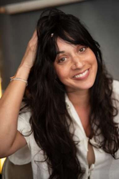 Smiling woman with long dark hair, hand resting on her head, wearing a white blouse indoors.