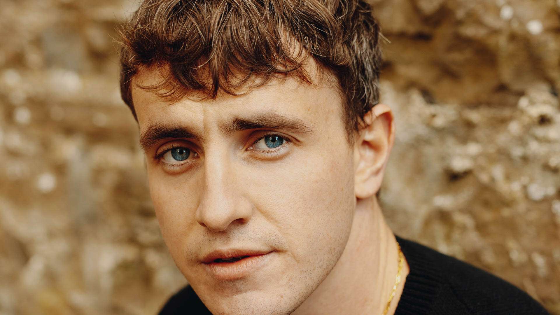 Close-up of a young man with blue eyes and brown hair against a stone wall backdrop.