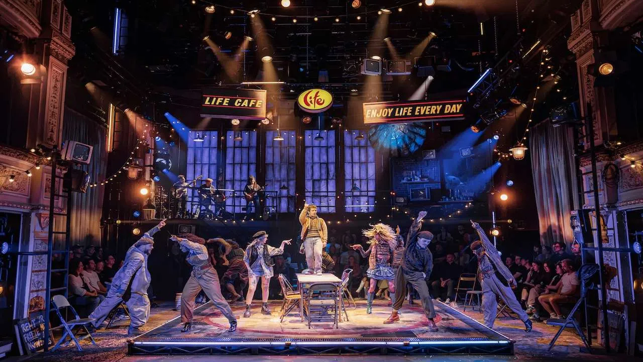 Energetic live-stage cabaret: dancers circle a central table while a singer stands on it; a band performs on a balcony as the audience watches.