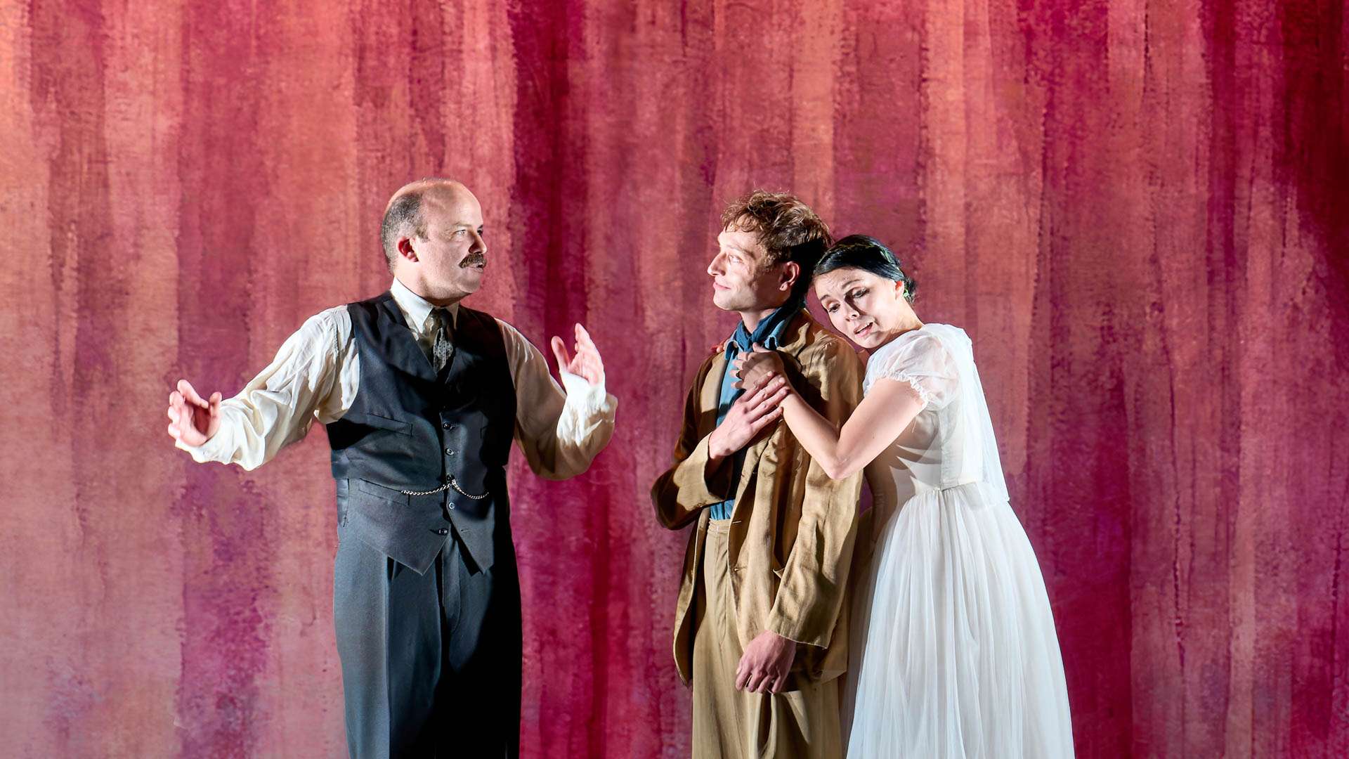 Stage scene with three actors in period costume: left man in a vest speaks, center man in tan smiles, right woman in white embraces him against a pink curtain backdrop.