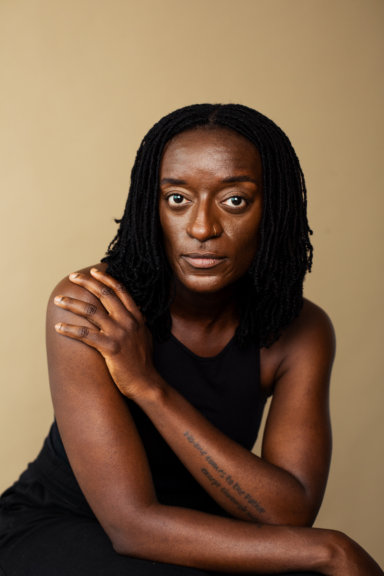 Portrait of a Black woman with dreadlocks wearing a black sleeveless top, posing with crossed arms against a beige background.