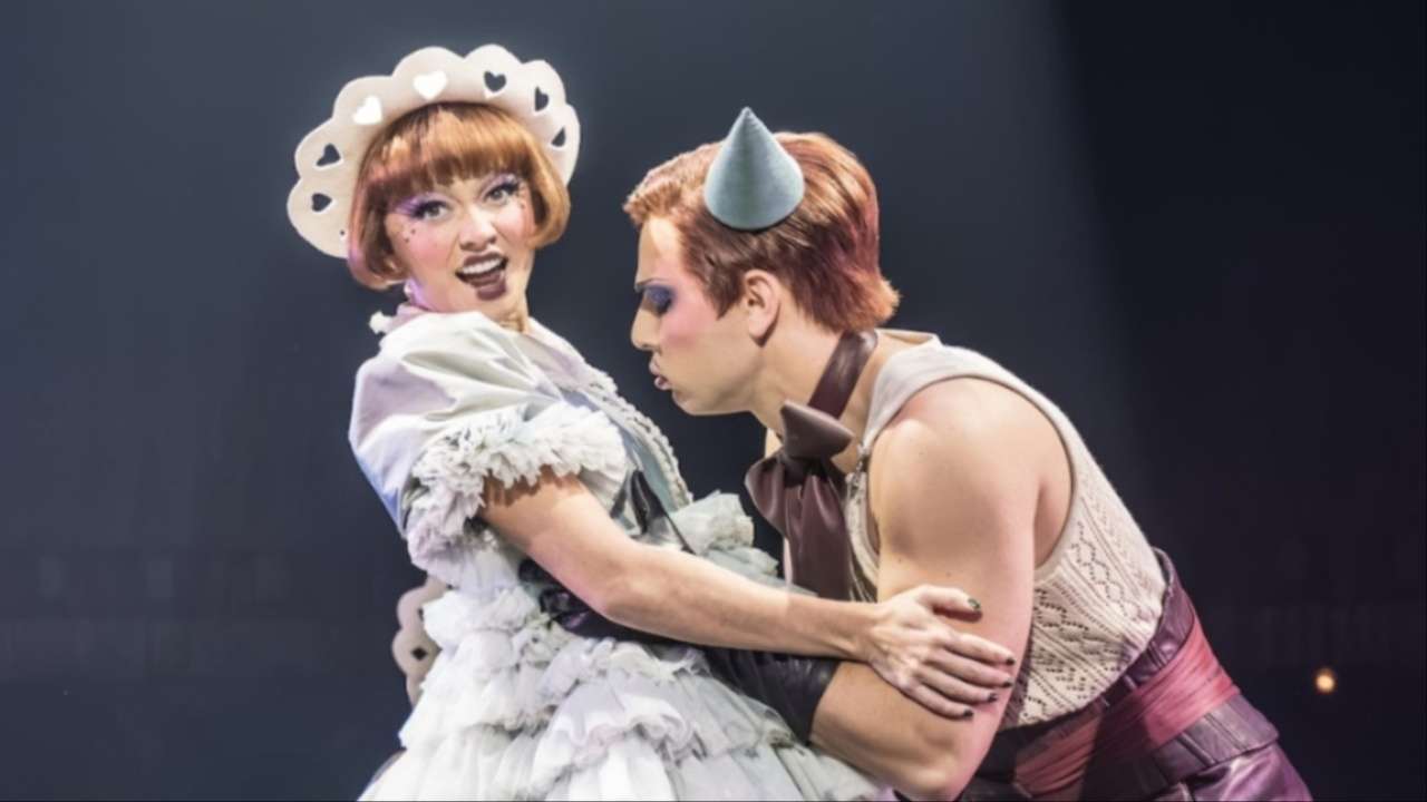 Two performers in elaborate stage costumes share a close heart-adorned headpiece facing the audience as the other actor leans in.