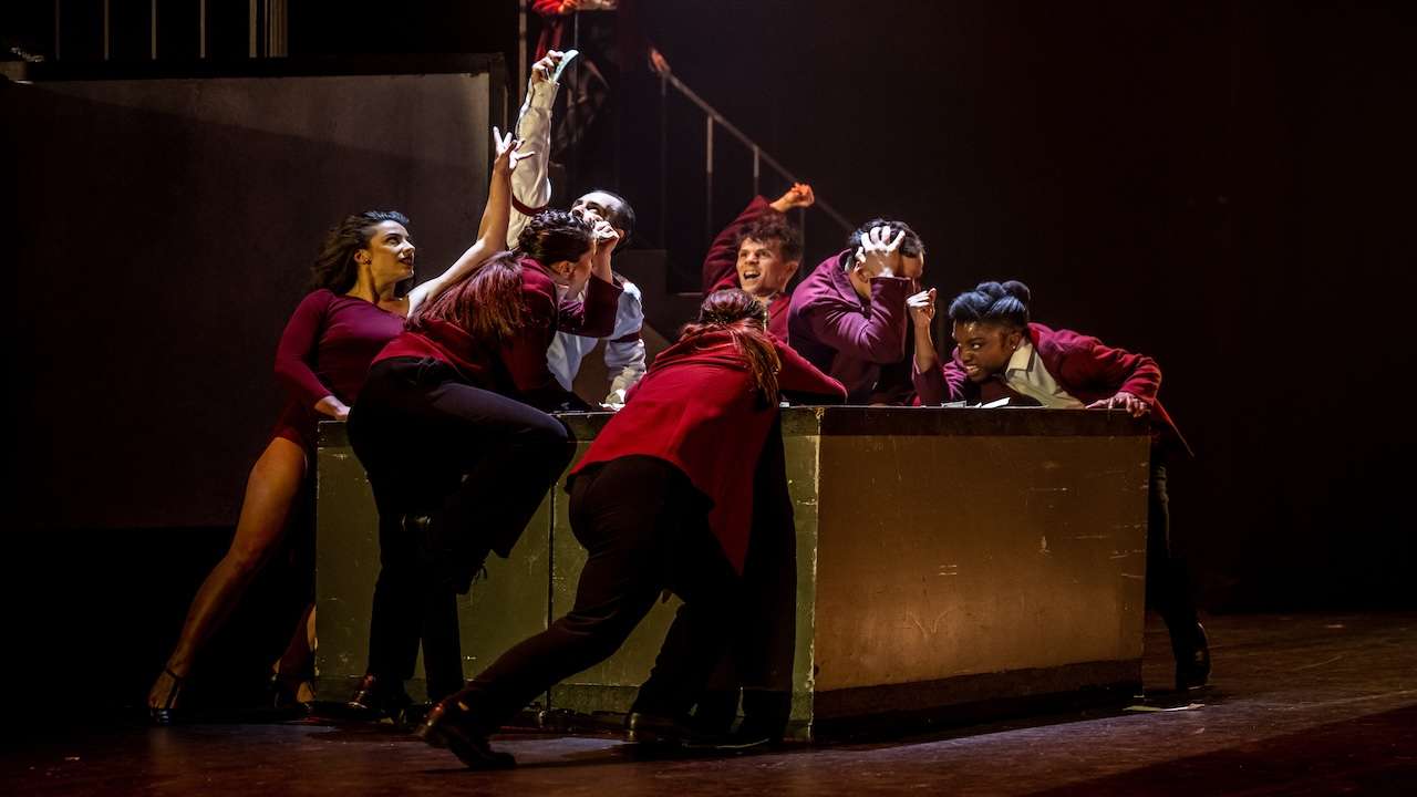 A group of dancers in burgundy costumes crowd around a large metal crate on a dark stage, reaching upward with intense expressions.