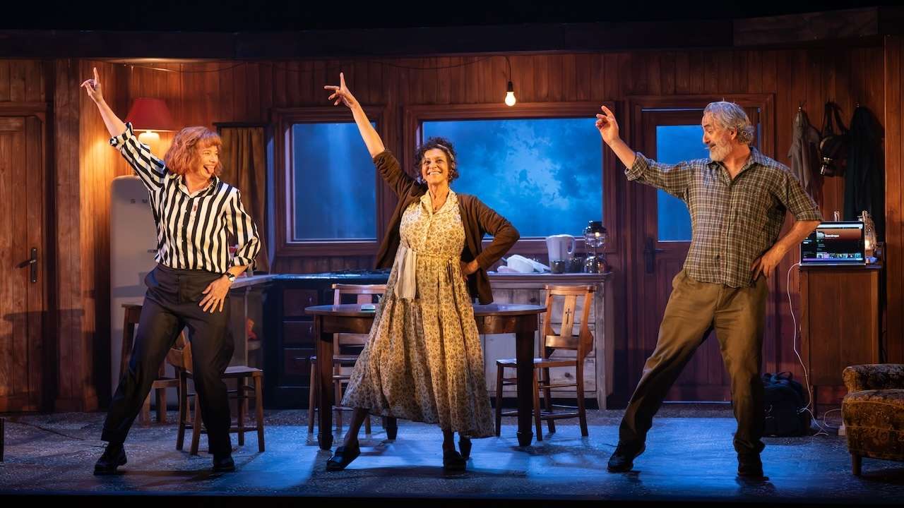 Three seniors dance on a wood-paneled stage, each with one arm raised in a pose at center stage.