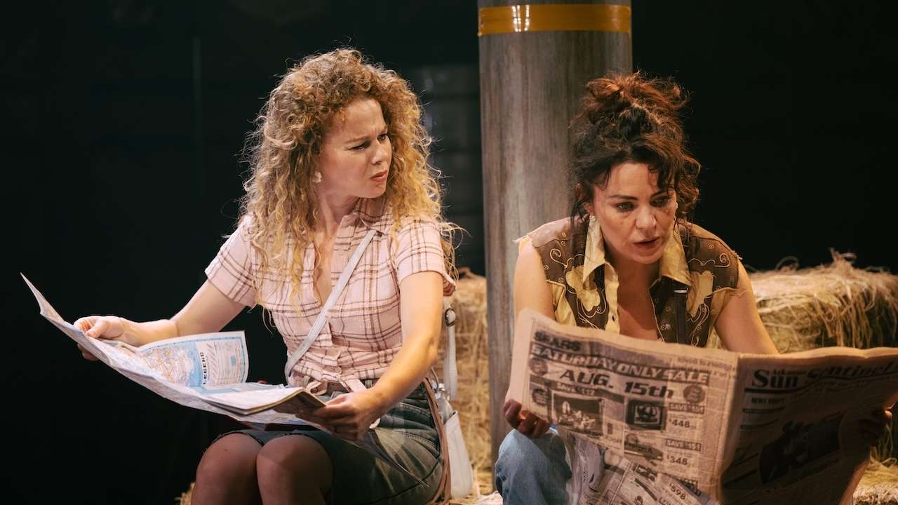 Two women sit on hay bales reading newspapers, on a dim stage, looking surprised or focused.