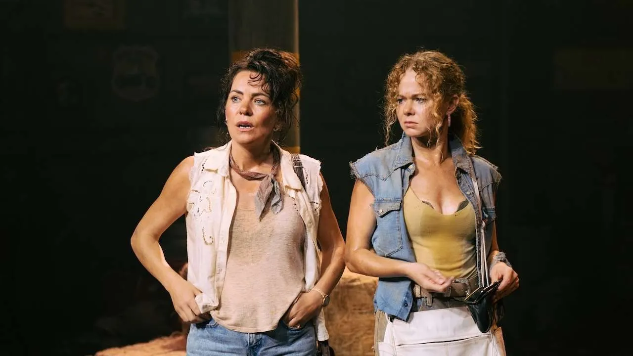 Two women on a dim stage, wearing rugged sleeveless tops and denim vests, looking concerned or tense during a performance.