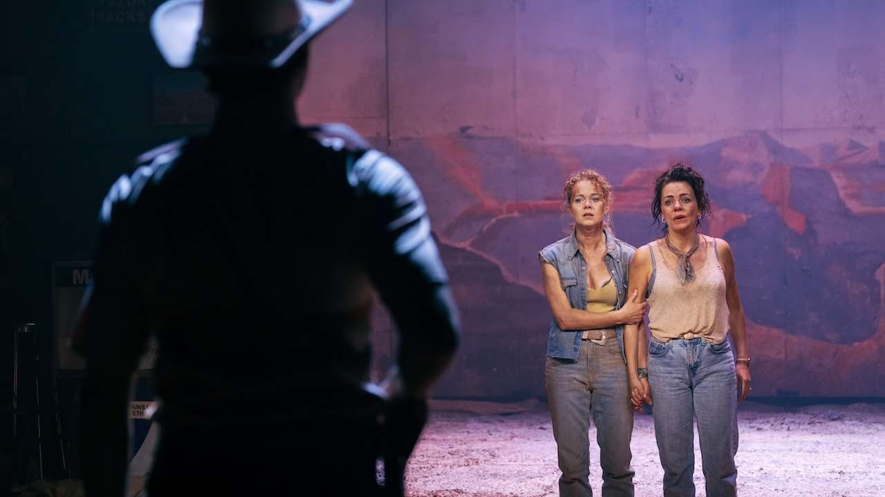 Two women stand on a dim stage, holding hands, while a man in silhouette watches from the foreground.