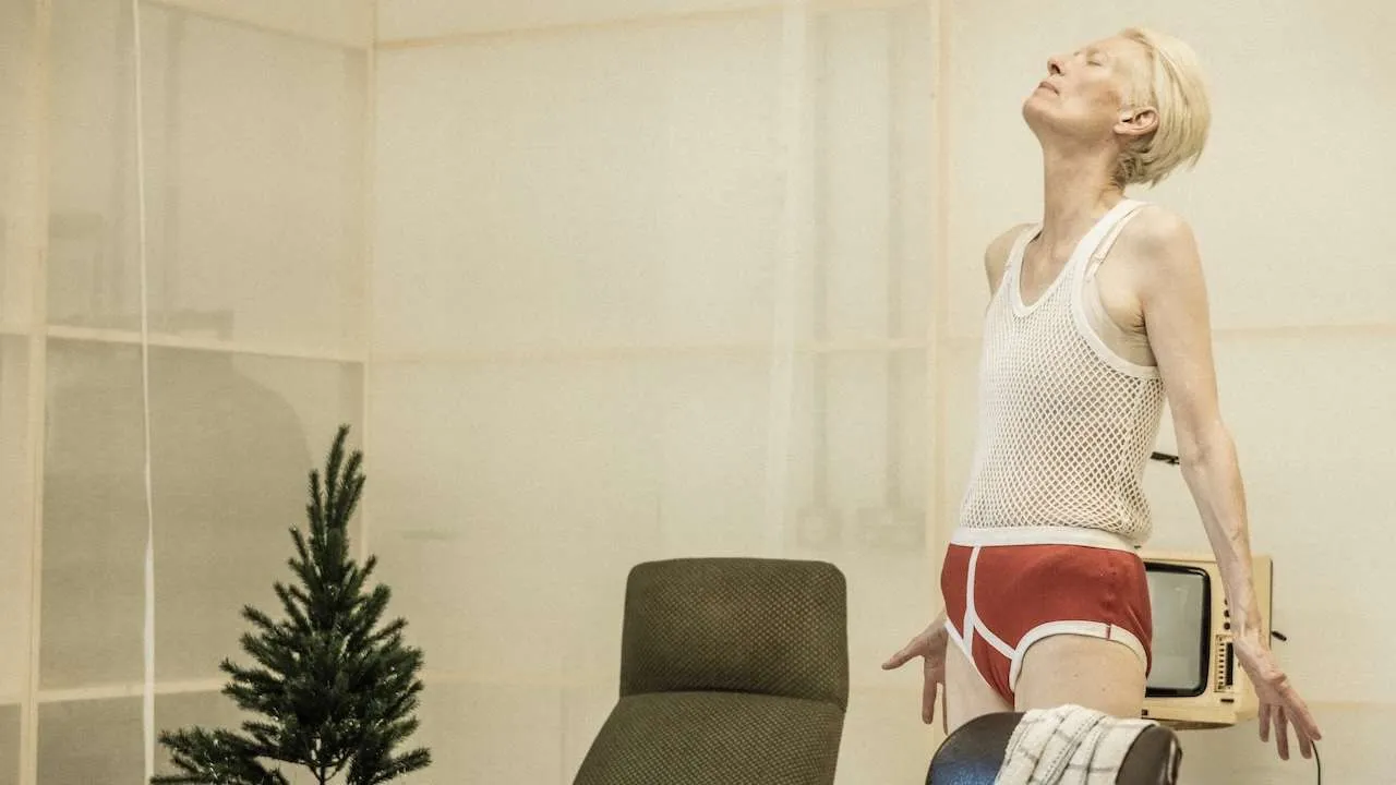 Woman in a white mesh top and red shorts stretches back in an office; a small Christmas tree and a vintage TV sit nearby.
