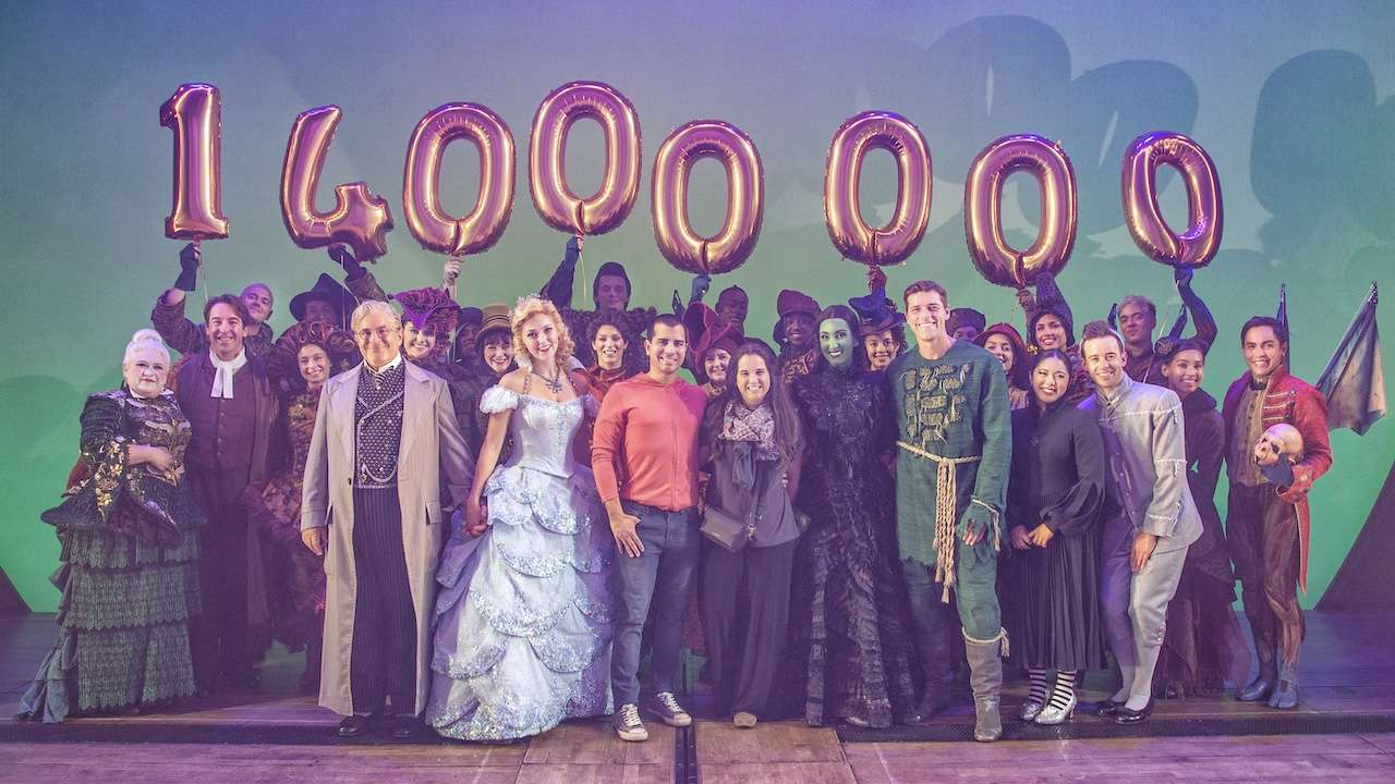 Group of stage performers in colorful period costumes posing with pink balloons reading 1,400,000 on a theater stage surface.