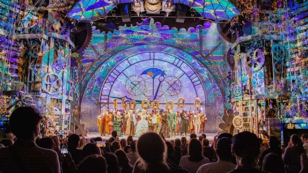 Colorful stage finale with a large clock backdrop, steampunk gears, and a line of performers bowing; audience watches from the foreground.