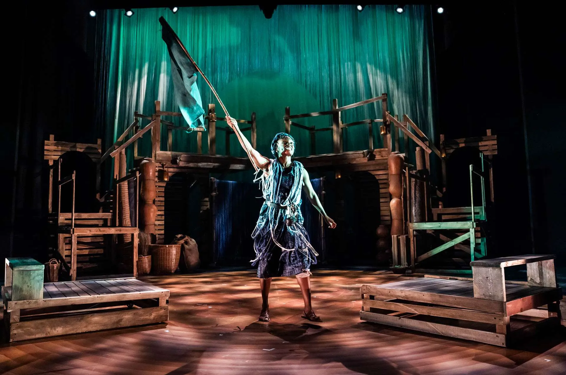 Performer in a blue shredded costume waves a flag on a wooden stage set with scaffolding and green backdrop at a theater production.
