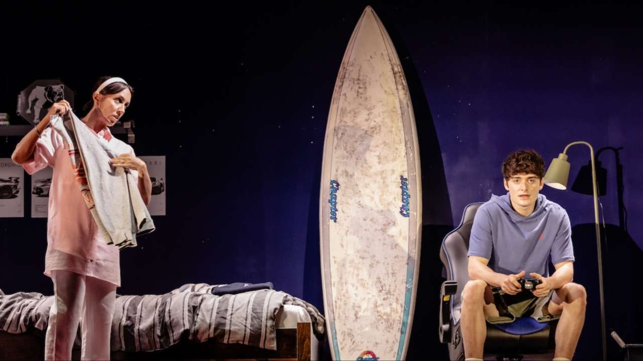 A woman folds laundry beside a teenage boy sitting in a gaming chair with a controller, a surfboard standing between them in a bedroom set.