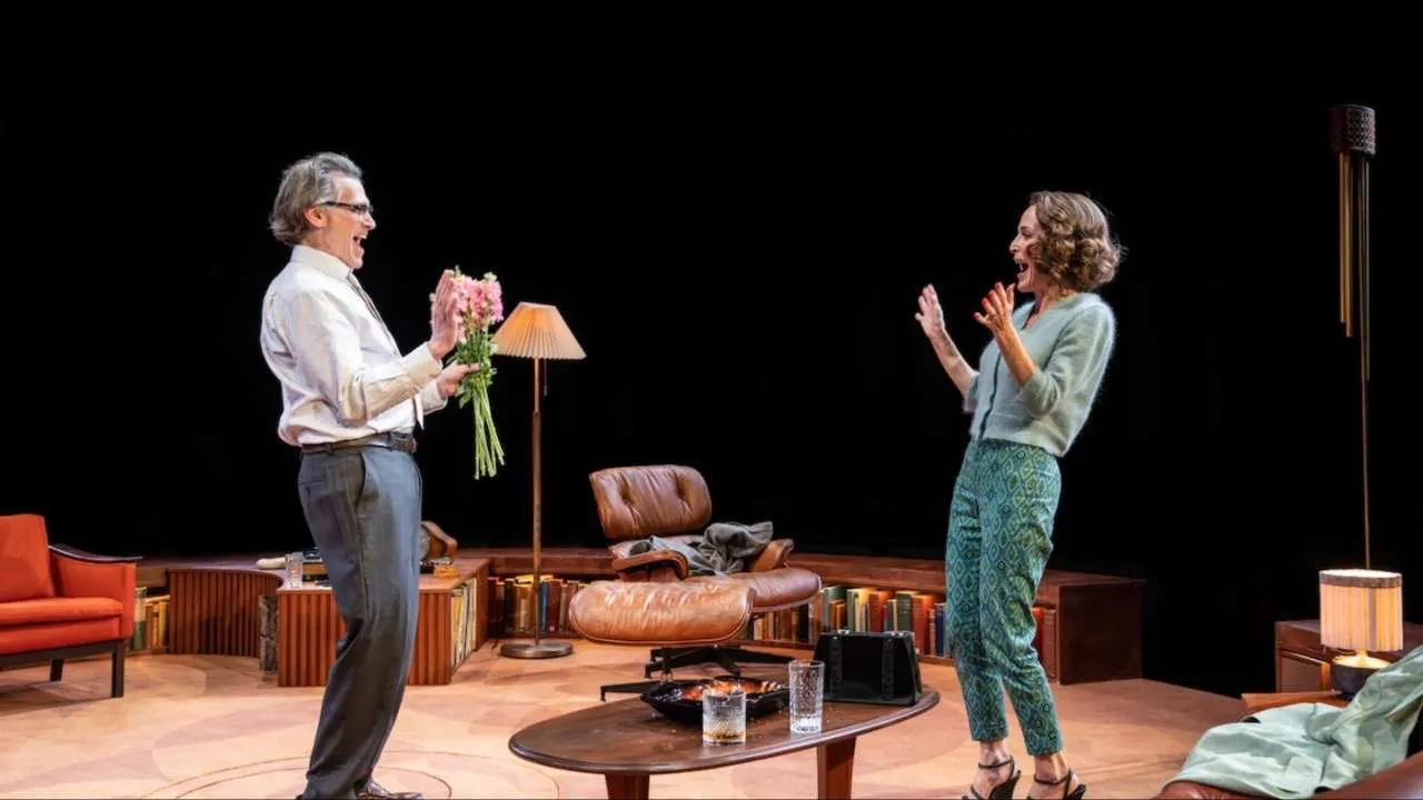 Older man hands pink bouquet to smiling woman on a theater stage set with mid-century furniture and warm lighting, as they laugh together.