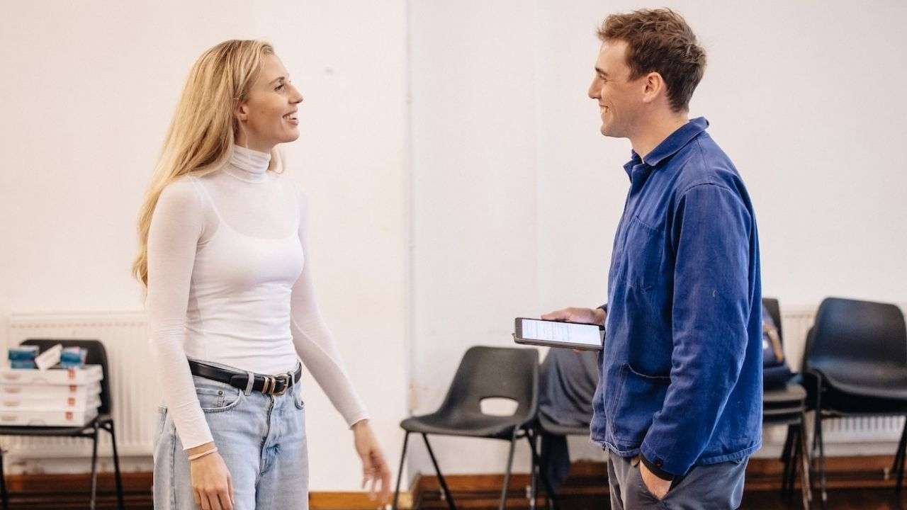 Two coworkers smiling and talking in a bright room; woman in a white turtleneck and jeans, man in a blue jacket holding a tablet.