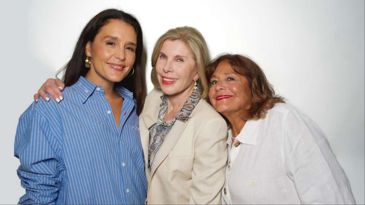 Three women stand close together smiling for a portrait, each with an arm around another in a friendly pose.