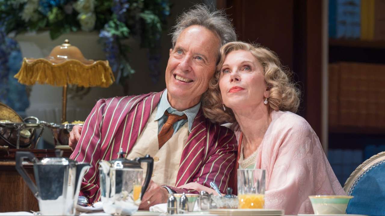 An older couple smiles, leaning close together at a dining table with silverware and a glass of orange juice, dressed in vintage style.