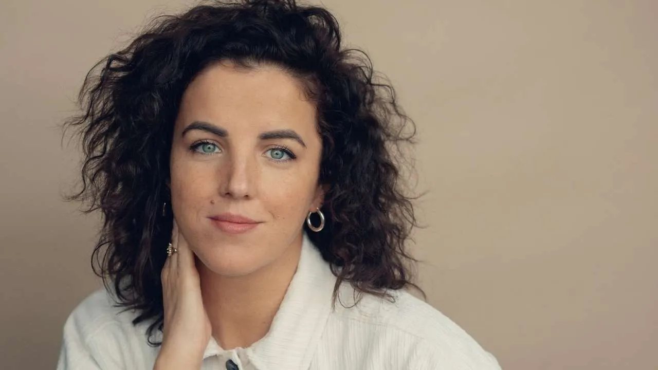 Portrait of a woman with curly dark hair and blue eyes in a white shirt against a beige background.