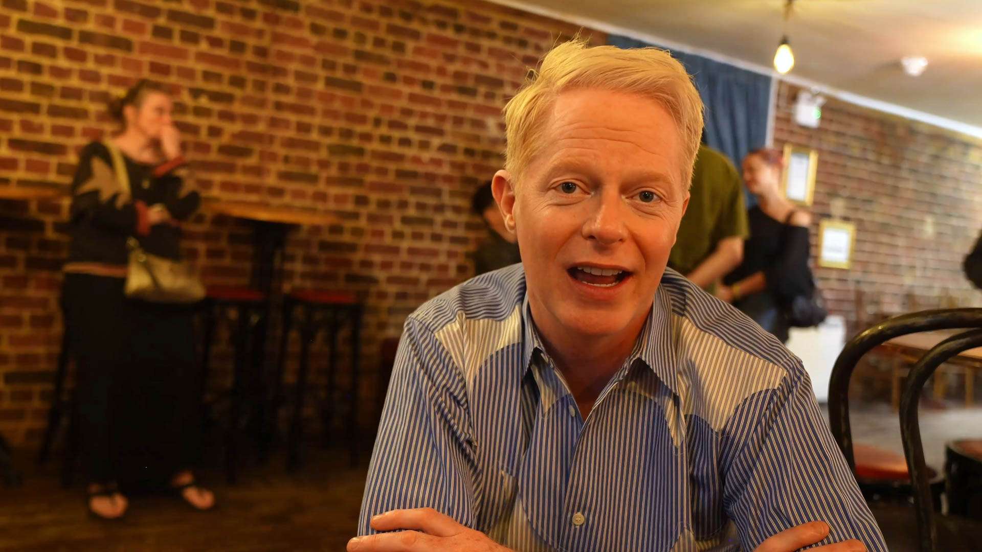 Man with blond hair in a striped shirt smiles at the camera in a brick-walled cafe, with blurred customers in the background.