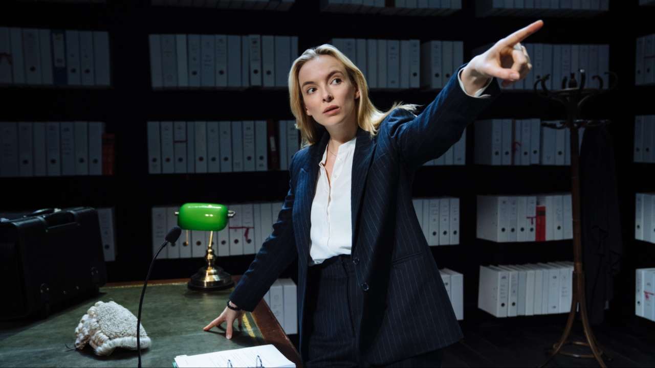 Jodie Comer in character as a barrister, pointing dramatically in a law chambers set lined with box files, with a barrister's wig resting on the desk.