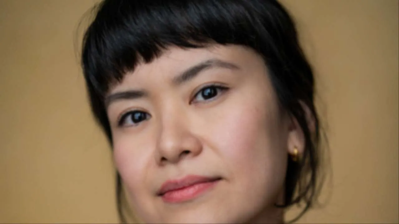 Close-up portrait of a woman with short black hair and subtle makeup, facing the camera against a plain beige background.