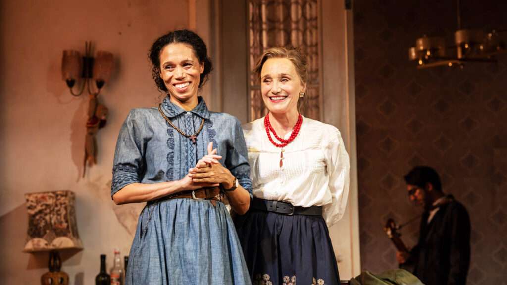 Two women in vintage dresses smiling on a stage, with a musician in the background.