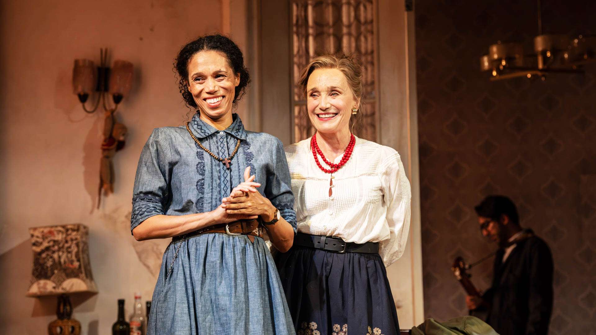 Two women in vintage dresses smiling on a stage, with a musician in the background.