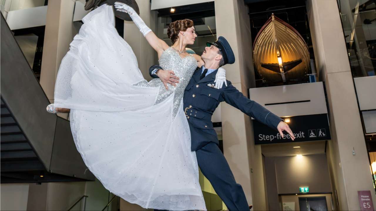 A ballet dancer in a sparkling white gown is lifted mid-pose by a dancer in RAF uniform inside the Imperial War Museum, with a lifeboat exhibit visible behind them