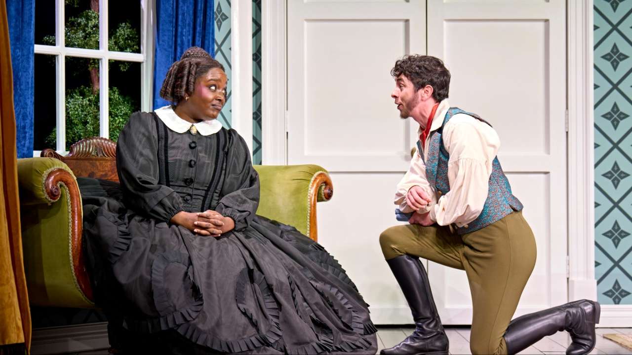 Woman in a black Victorian gown sits on a green sofa as a man in a vest and boots kneels and speaks to her in a period interior.
