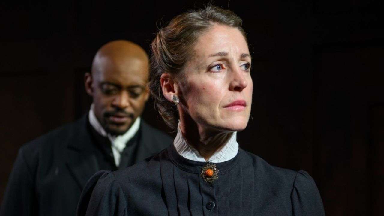 Woman in a Victorian-style black dress with a white high collar and orange brooch looks to the right; a man in a dark suit stands behind her on stage.