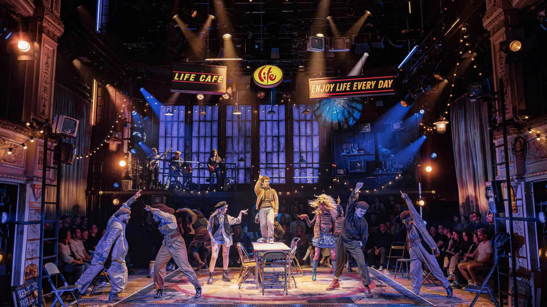 Energetic stage show with a center-stage singer, dancers in front, and a live band on a balcony, under warm lights and banners that read “Life Cafe” and “Enjoy Life Every Day.”