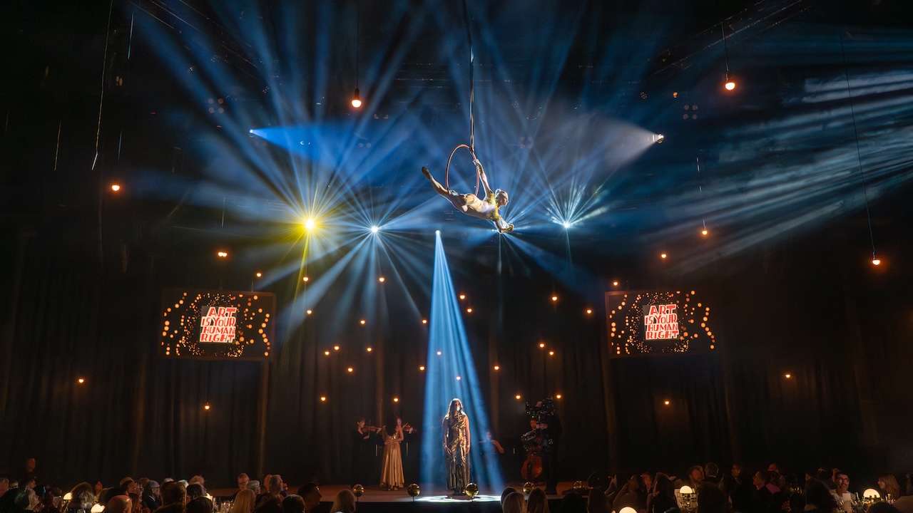 Aerialist performs on a hoop suspended from above, bathed in blue and yellow spotlights over a dark theater and seated audience below.