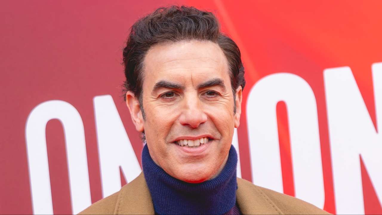 Sacha Baron Cohen smiling at the camera against a red background at an event.
