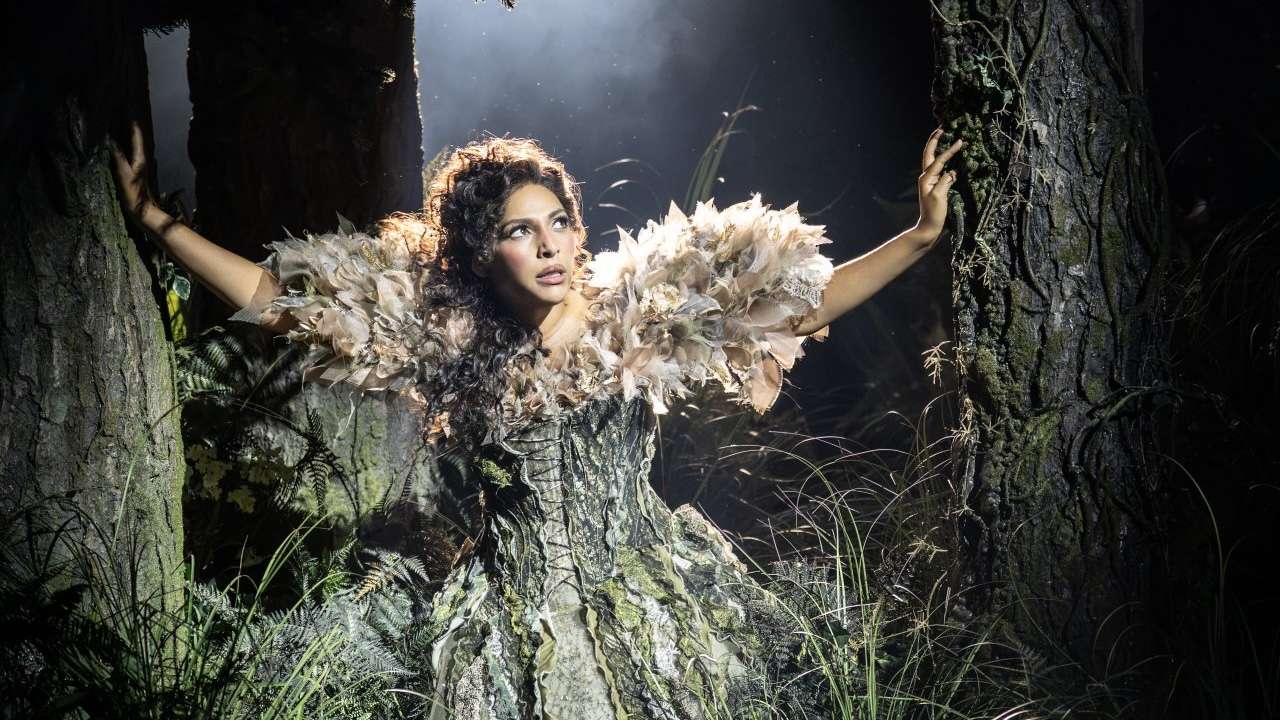 Woman in a feathered gown stands between two trees in a dark forest, arms extended, looking upward.