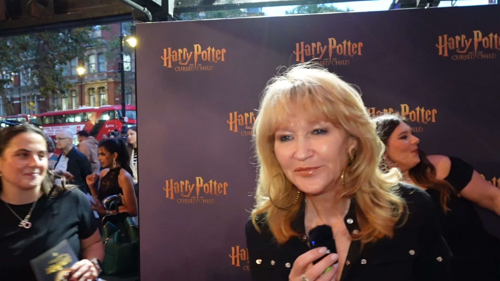 Blonde woman being interviewed in front of a Harry Potter: Cursed Child backdrop, with fans and a red bus visible in the background.