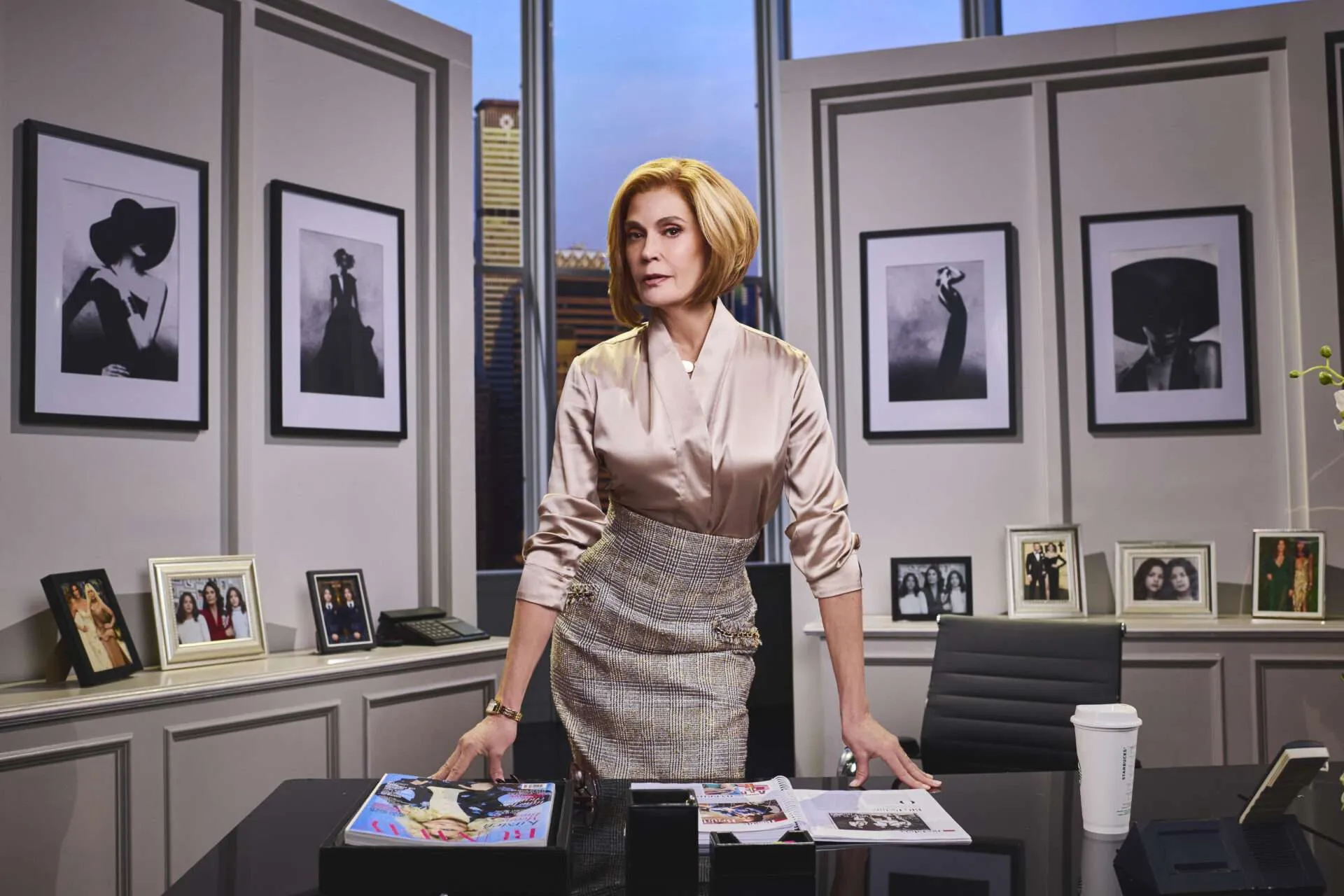 Businesswoman standing behind a desk in a modern office, surrounded by framed photos on the walls and desk items including magazines and a take-away coffee cup.