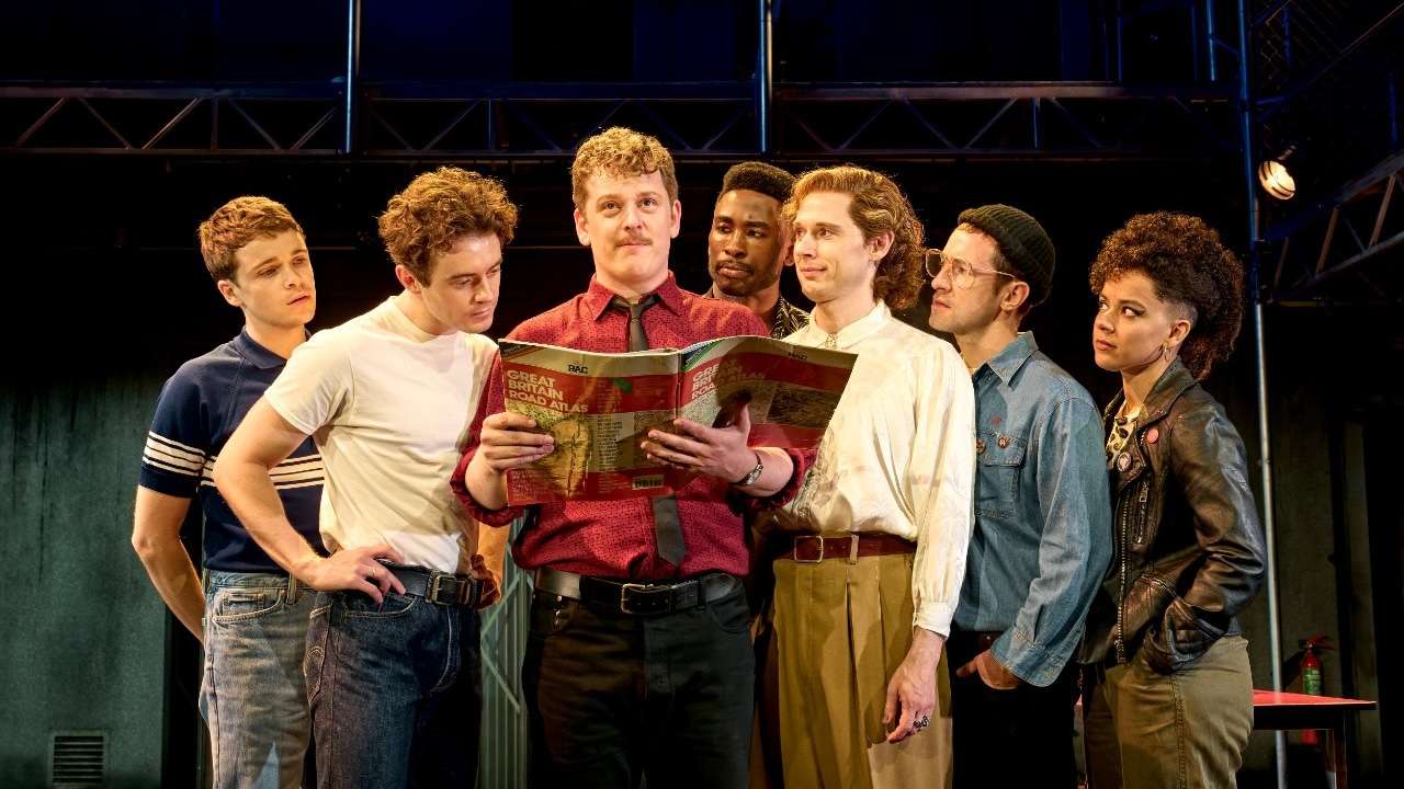 Group of seven actors on stage gathered around a map, reading it together during rehearsal.