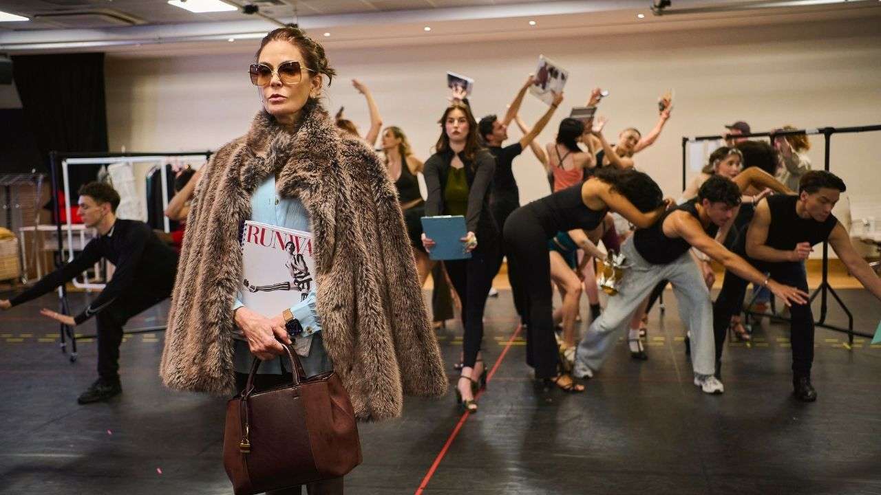 Teri Hatcher in oversized sunglasses and a fuzzy brown coat holds a leather handbag while posing in the foreground, with ensemble cast members performing choreography with raised arms in a rehearsal studio behind her.