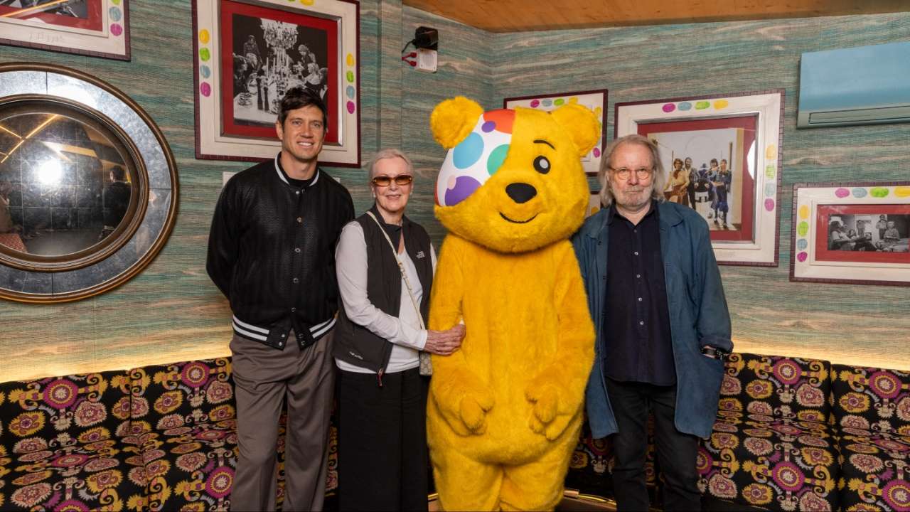 Two adults and a woman pose with a yellow bear mascot in a colorful, retro-style lounge with framed photos on the wall and a patterned sofa behind them.
