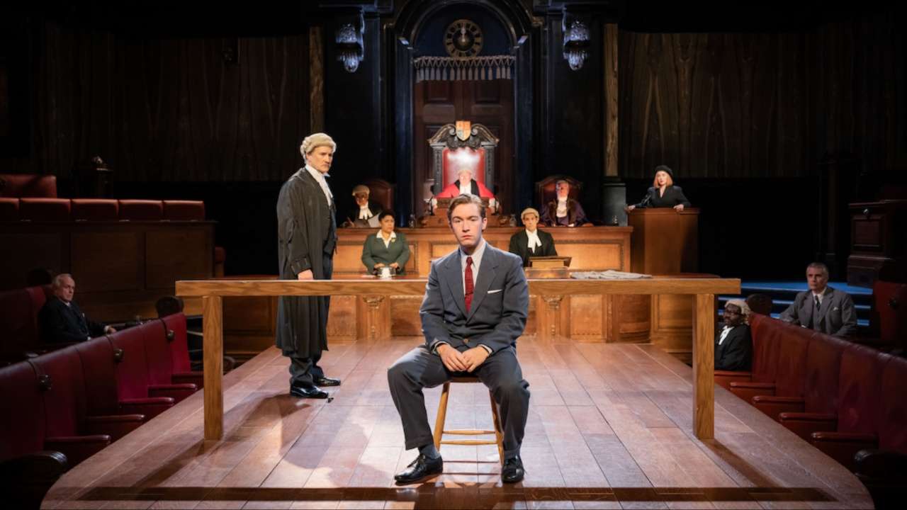 A man in a gray suit sits on a wooden chair at a courtroom stage, with a judge and legal staff behind a long bench and spectators around.
